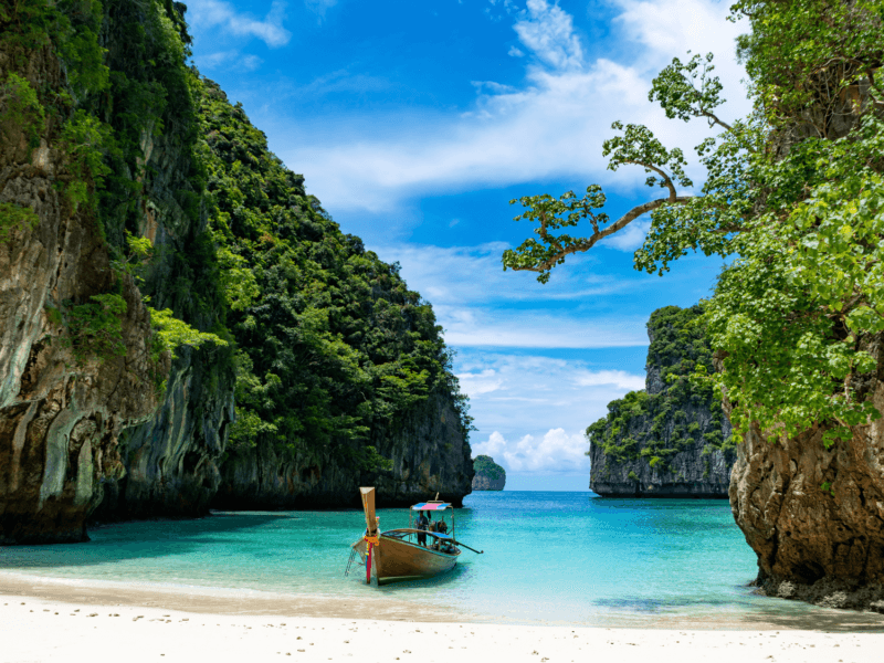 Hidden lagoon at Phi Phi Island featuring a traditional longtail boat, turquoise waters, lush green cliffs, and a serene blue sky.