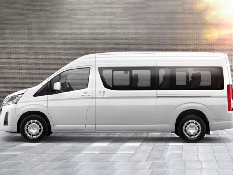 Side view of a white Toyota commuter van parked on a modern paved surface, ideal for airport transfers.