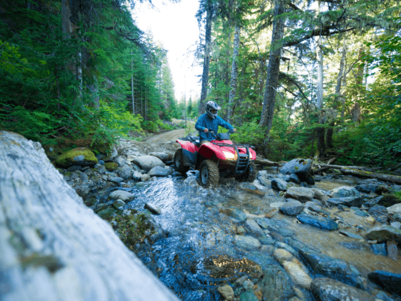 Adventure ATV rider crossing a shallow river through rugged terrain in Phuket's lush forest landscape.