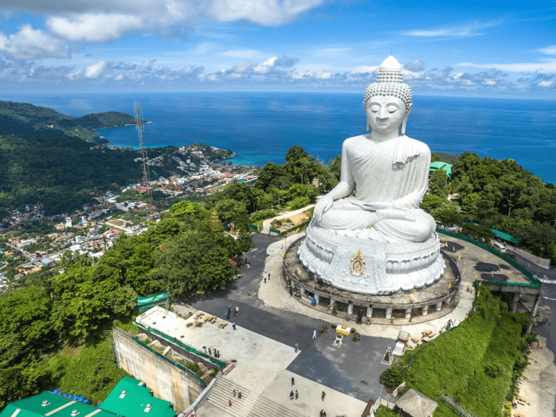 Aerial view of the Big Buddha statue in Phuket, Thailand, overlooking lush greenery and the Andaman Sea under a bright blue sky.