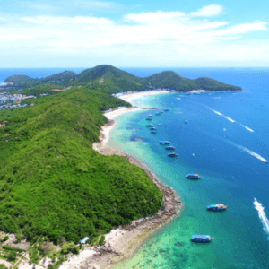 Coral Island Private Charter: Aerial view of Coral Island showcasing lush green hills, turquoise waters, and anchored boats, with the horizon stretching under a clear blue sky.