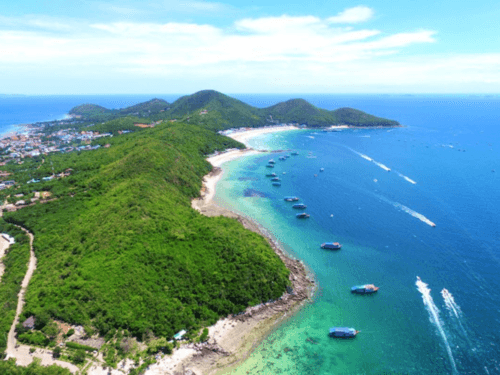 Coral Island Private Charter: Aerial view of Coral Island showcasing lush green hills, turquoise waters, and anchored boats, with the horizon stretching under a clear blue sky.