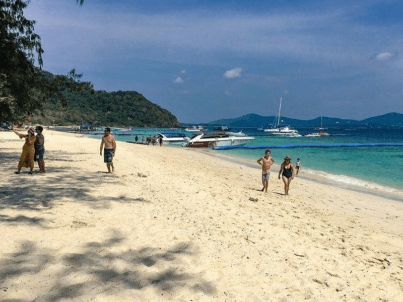 Visitors strolling along Coral Island's sandy beach with speedboats anchored in the turquoise water and lush green hills in the background. Perfect for family getaways and island tours.