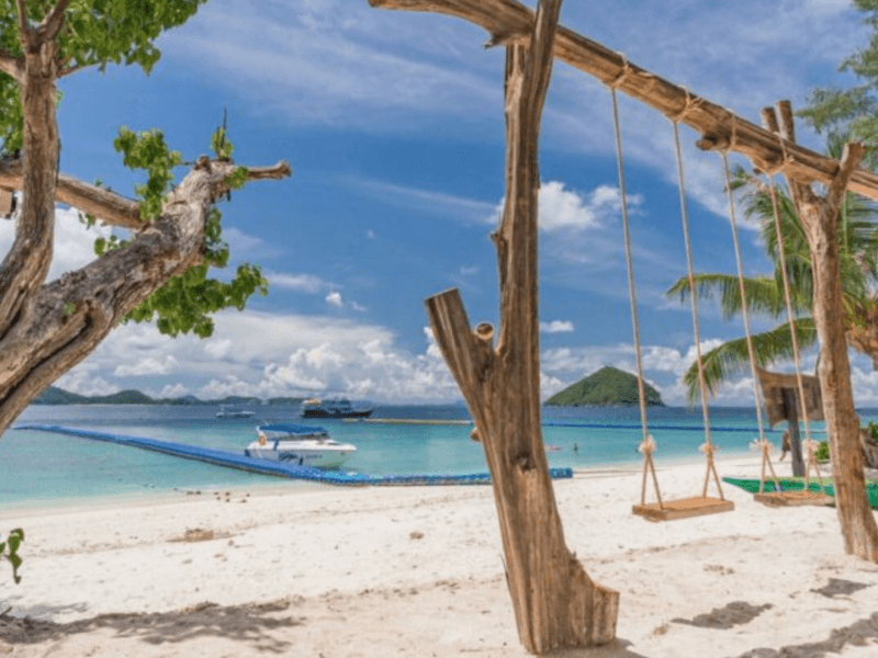 A rustic wooden swing overlooking Coral Island's white sandy beach and crystal-clear waters, with speedboats and a scenic island in the background under a sunny sky.