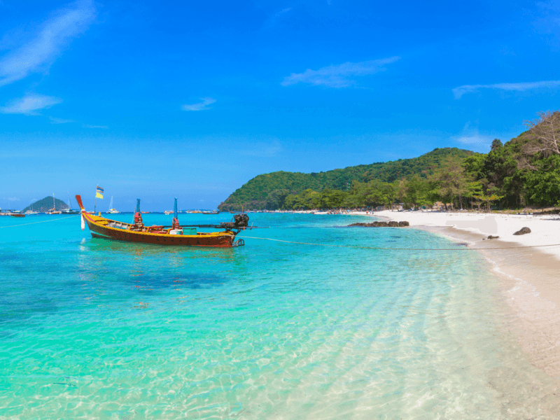 Coral Island tour: Crystal-clear waters with a traditional longtail boat at Coral Island, Phuket, Thailand, surrounded by lush greenery and blue skies.