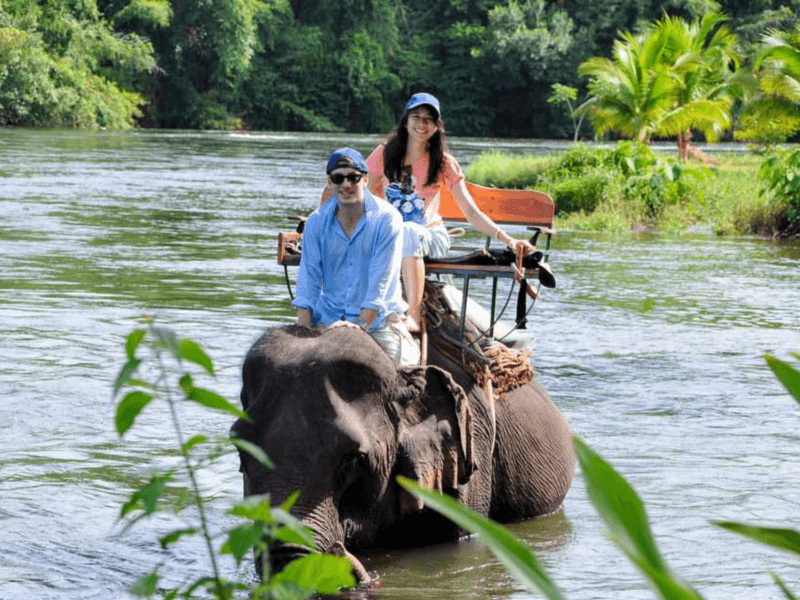 Couple enjoying a serene elephant trekking experience along a picturesque river in Phuket, surrounded by lush greenery and tropical scenery.