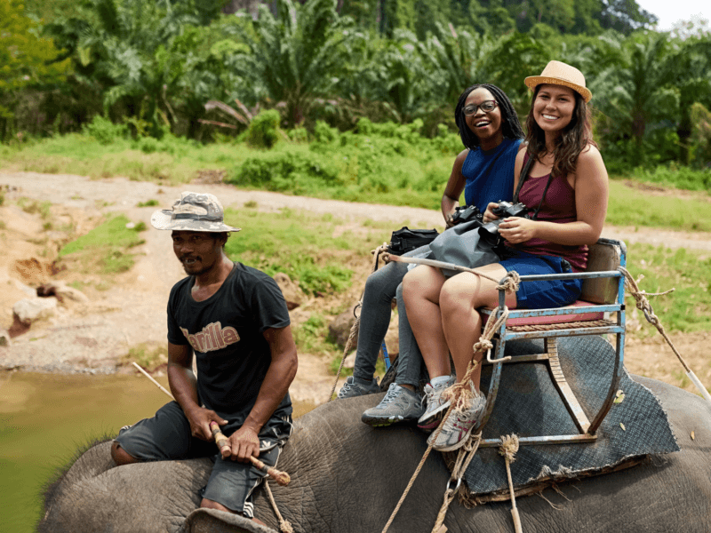 Two women enjoying an elephant trekking tour in Phuket with a guide in a lush natural setting.