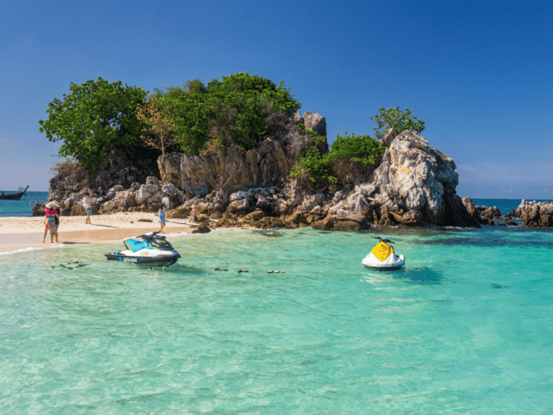 Tourists enjoying a sunny day on Khai Island with jet skis floating on the turquoise waters near the rocky shore.