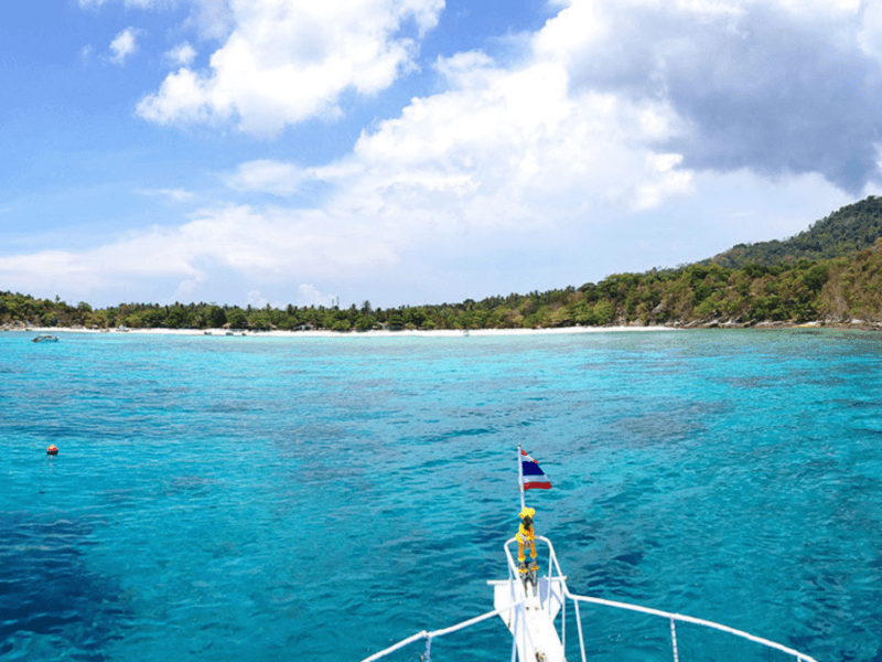 Scenic view from the boat's bow overlooking the pristine turquoise waters and lush shoreline of Khai Island under a partly cloudy blue sky.