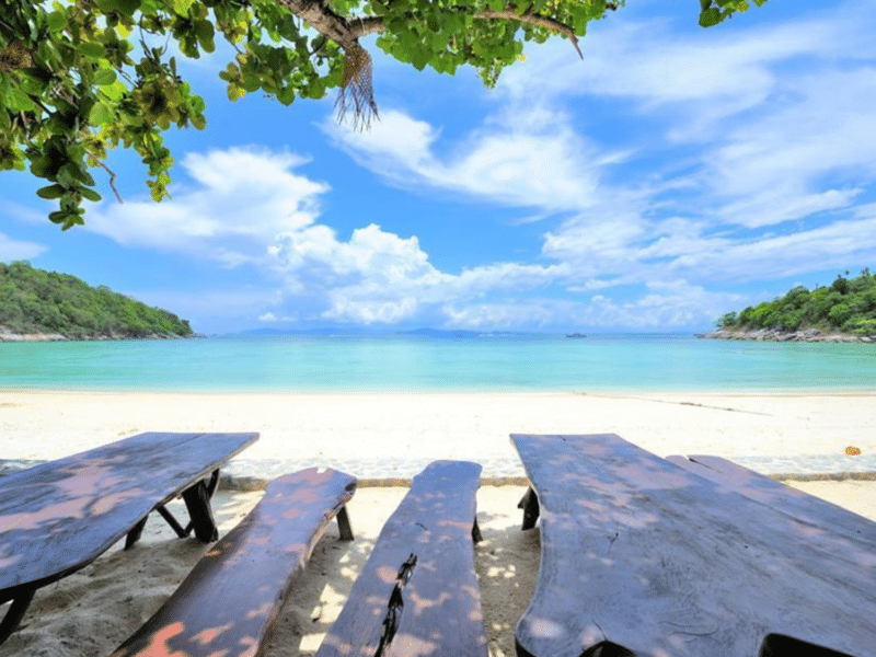 Wooden picnic benches overlooking the tranquil turquoise waters and white sandy beach of Khai Island, framed by lush greenery.