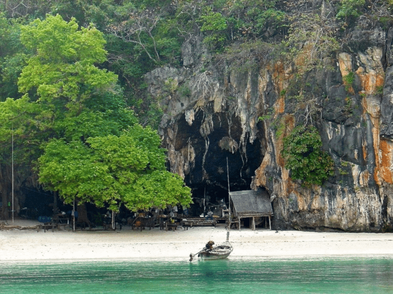 A scenic beachfront at Koh Yao Yai with lush green trees, a limestone cave, and a small wooden hut by the shore.