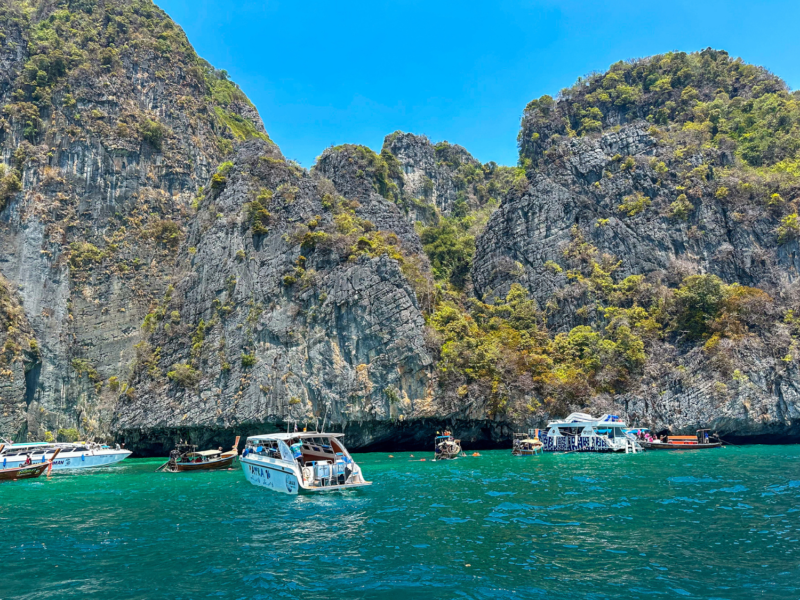 Speedboats and longtail boats anchored near the towering limestone cliffs of Phi Phi Islands under a clear blue sky.