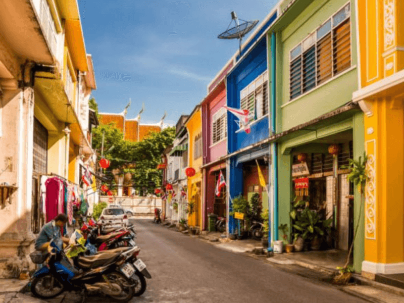 A vibrant street in Phuket Old Town with colourful colonial-style buildings and parked motorbikes under a bright sky.