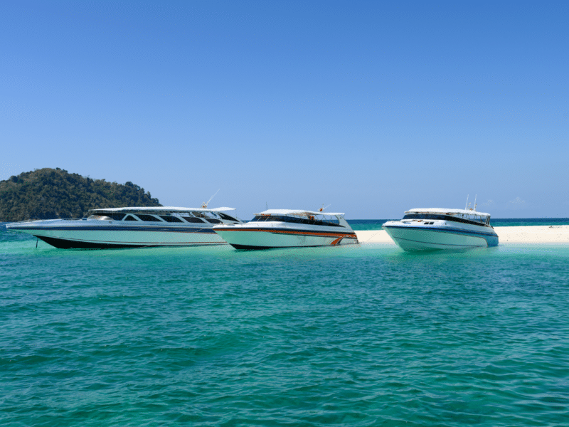 Three private speedboats anchored on a pristine tropical beach with turquoise waters and a lush island in the background.