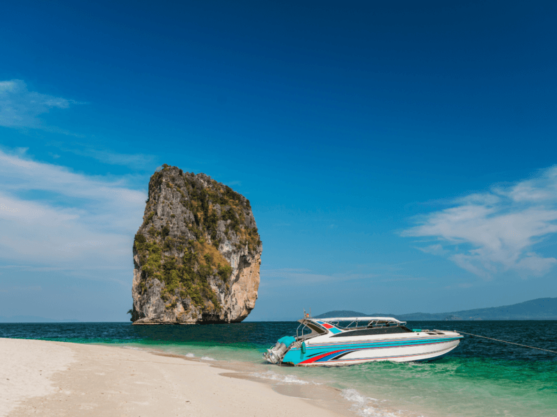 A private speedboat anchored on a pristine white sand beach with a towering limestone island in the background at Phi Phi Islands.