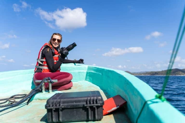 adventure charter in phuket: Smiling marine biologist holding a professional camera, seated on a boat during a sunny day at sea.